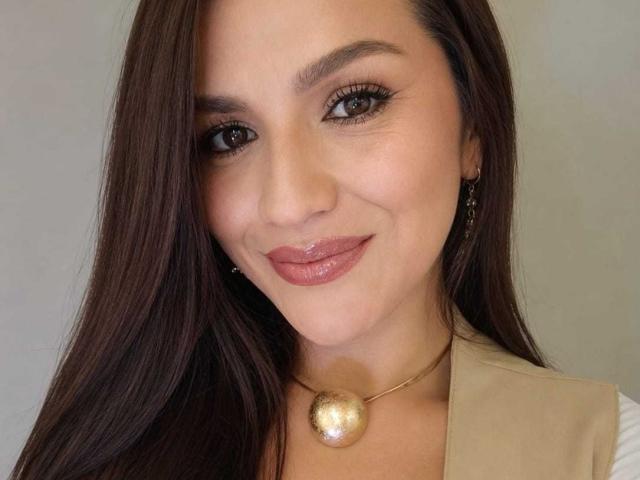 CameronViviane has shiny brown eyes, long straight brown hair, glossy pink lips, and a gold ball necklace. She's smiling softly with a tan vest.