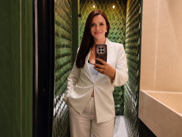 CameronViviane with long brown hair, wearing a white blazer and white pants, holding a phone, smiling, showing a subtle cleavage, standing in a green-walled corridor.