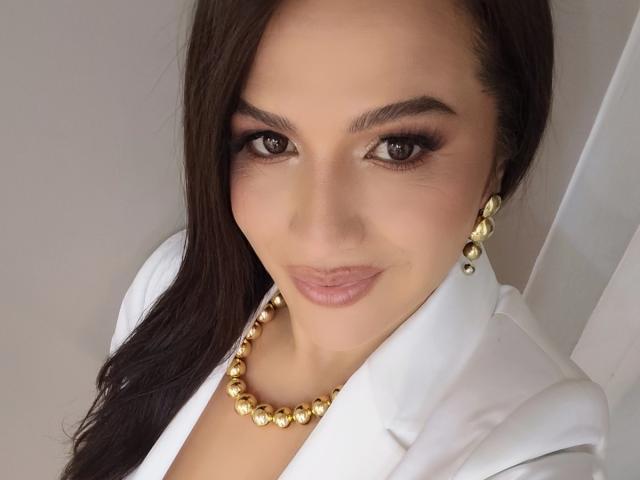CameronViviane with dark brown hair, wearing a white jacket, gold earrings, and a gold bead necklace, smiling with makeup and lipstick.