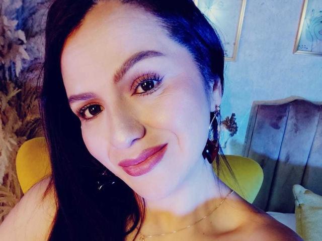 CameronViviane has dark hair, blue eyes, and pink lipstick. She's smiling with a subtle nose and wearing dangling earrings. Her bare shoulders are visible.