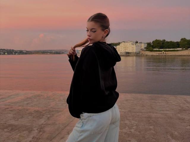 BethanySandhoff with a slim ass, wearing a black hoodie and white pants, standing by a river at sunset, holding her brown hair with a calm expression.