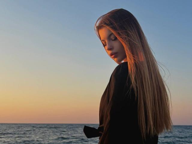 BethanySandhoff with long, straight brown hair, wearing a black shirt, looking down with a soft, pensive expression, standing by a sunset over the ocean.