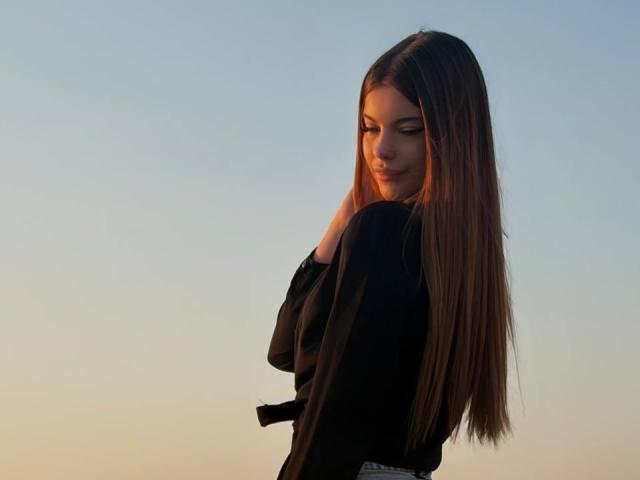 BethanySandhoff with long brown hair, black shirt, and subtle smile, looking over her shoulder, hand in hair, soft sunset light, clear blue sky, gentle evening glow.