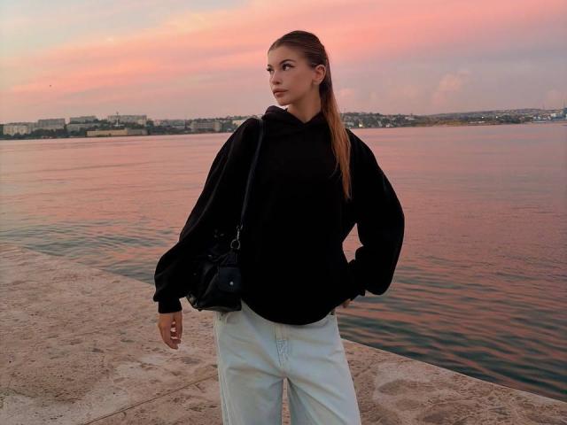 BethanySandhoff with long brown hair, black hoodie, white pants, black crossbody bag, standing by a calm water, pink-orange sunset sky, hands in pockets, looking to the side.