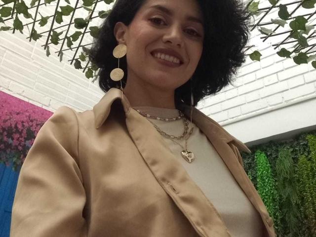 LilaaSalmon with curly black hair, smiling, wearing a beige jacket, white top, gold earrings, and necklace, showing her tits slightly under the jacket. Outdoor, green leafy background.
