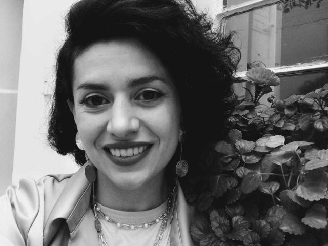 Black-and-white photo of LilaaSalmon with curly dark hair, smiling, wearing a shiny jacket, necklace, and earrings, standing next to a leafy plant, showing her teeth.