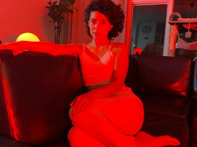 LilaaSalmon with curly hair, red-lit, sitting on black leather couch, wearing sheer white dress, legs crossed, arm on couch, looking serious, soft glow from round yellow lamp.