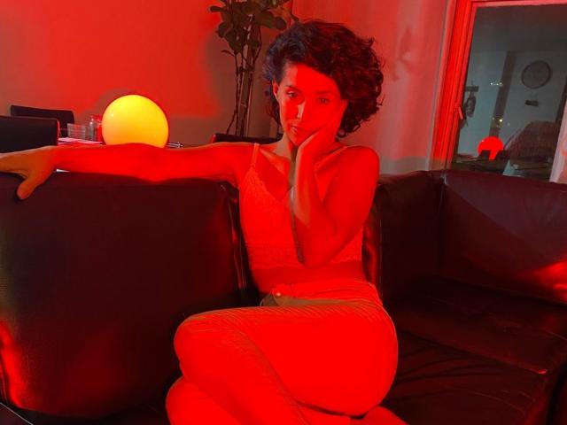LilaaSalmon with curly dark hair, red-lit skin, and a pensive expression, sits on a black leather couch, arm resting on a glowing orb, wearing a tight, sleeveless top.