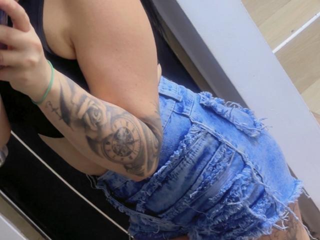Kkatalina's arm tattoo featuring a detailed, black-and-grey owl with a compass, wearing frayed, blue, denim shorts and a black top, showing her toned, tan, side profile.