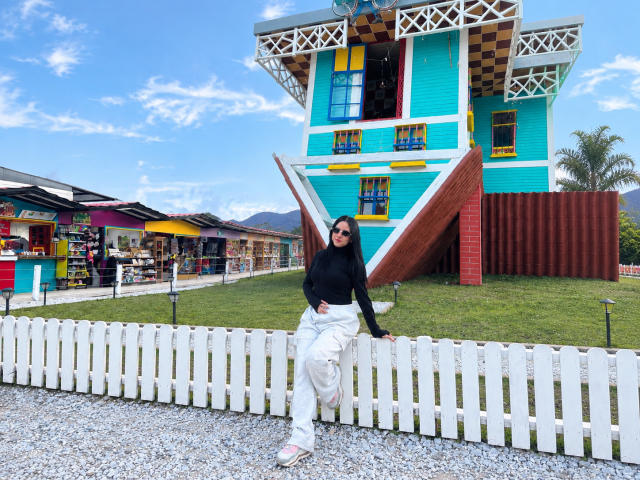 MiaAylen with long black hair, black top, white pants, and pink sandals, leaning on white picket fence, standing in front of colorful, geometric building, blue sky above.