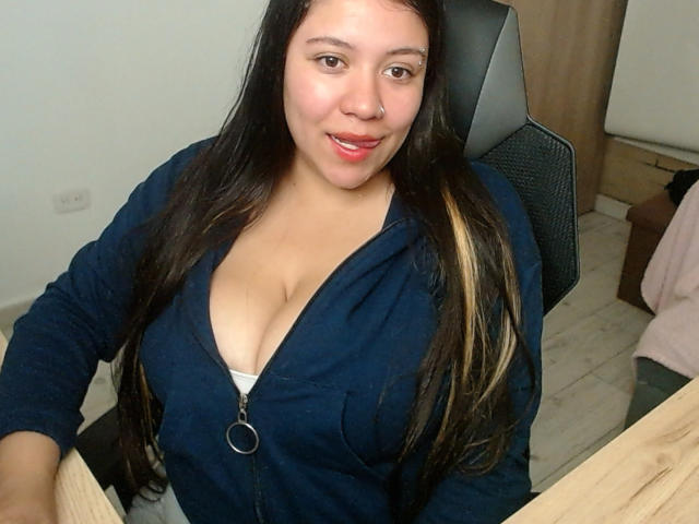 KrisGarner with long black hair, wearing a navy zip-up hoodie, revealing white bra and cleavage, sitting in a chair, smiling at the camera, in a simple room.