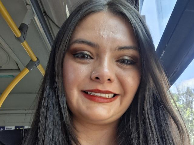 KatherinLatin a les cheveux longs et bruns foncés, un petit piercing au nez, et elle sourit avec du rouge à lèvres, montrant ses dents. Elle est à l'intérieur d'un bus avec des poteaux jaunes..