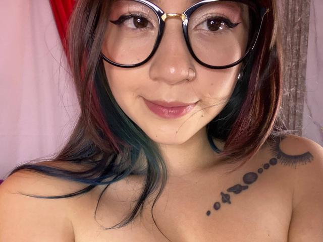 EmmaFeri with big glasses, brown eyes, nose ring, and smiling pink lips. She's topless, showing a tattoo of dots and eyelashes on her right shoulder.