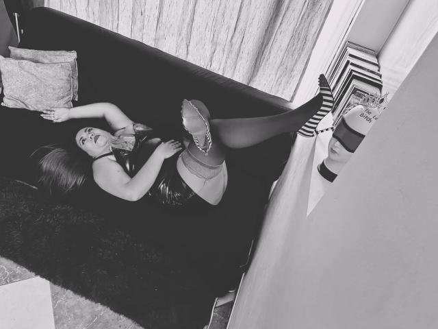 LexyDom is lying on a black couch, legs up, wearing sheer stockings, striped heels, and a leather skirt, with her hand on her shiny boobs, in a black-and-white photo.