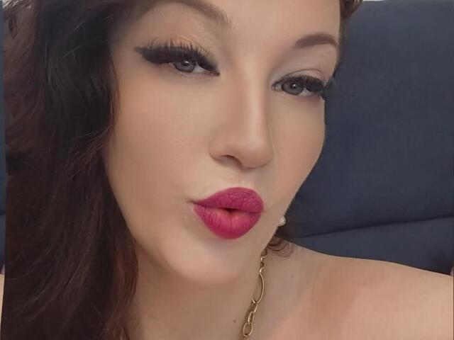 VanessaSquirtt has pouty pink lips, heavy black eyeliner, long dark eyelashes, and wavy brown hair. She's wearing a gold chain necklace and has a fair skin tone.