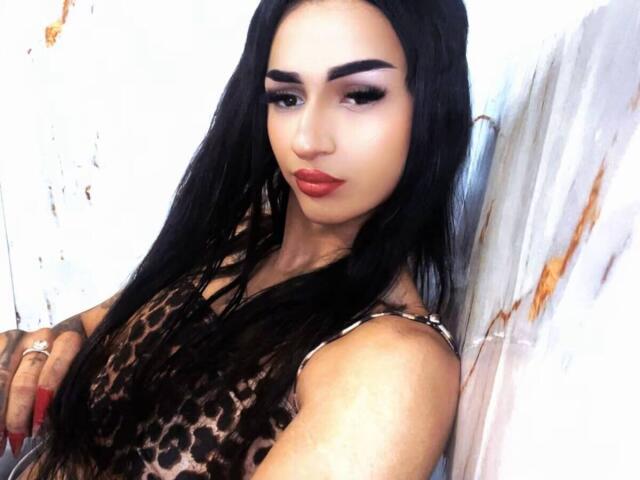 IrisIrs has long black hair, thick eyebrows, red lipstick, and wears a leopard print dress. She's leaning against a white wall with a slight smile.