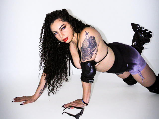 ScarlattaaDesire with curly black hair, red lipstick, black leather arm sleeves, black top, purple skirt, black boots, and tattoos, on all fours, looking at the camera.