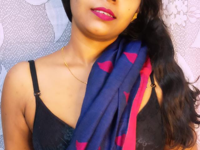SimplySoumi con lápiz labial rosa, un sujetador de encaje negro, un collar dorado y cabello negro largo, sosteniendo una bufanda azul con corazones rojos, cubriendo sus senos..