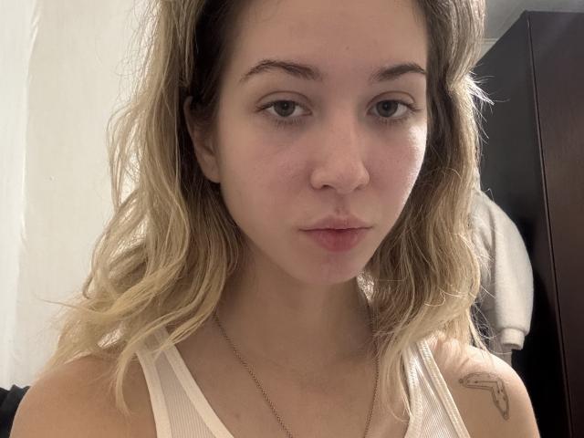 Leona Kuharik hat welliges blondes Haar, helle Haut und ein kleines Tattoo an ihrer rechten Schulter. Sie trägt ein weißes Tanktop mit einem Aufdruck von geschwollenen Lippen und eine dezente Halskette..
