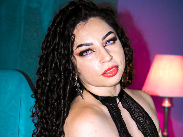 ScarlattaaDesire mit lockigem, schwarzem Haar, blauen Augen, rotem Lippenstift und einem schwarzen Top mit Halter, das ihre Brüste zeigt und ein süßes Nasenpiercing trägt. Sie wirkt verführerisch und selbstbewusst..