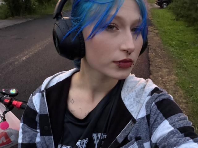 DarylSanks with blue hair, black headphones, nose ring, lip piercing, striped hoodie, and black shirt, holding a bike handle on a road.