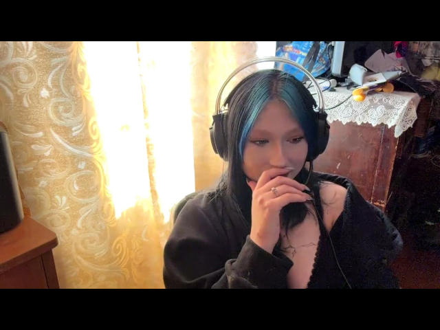 DarylSanks with blue highlights, wearing black headphones, has finger near mouth, black hoodie, pale skin, slight smile, in cozy room, wooden furniture, lace tablecloth, and TV in background.