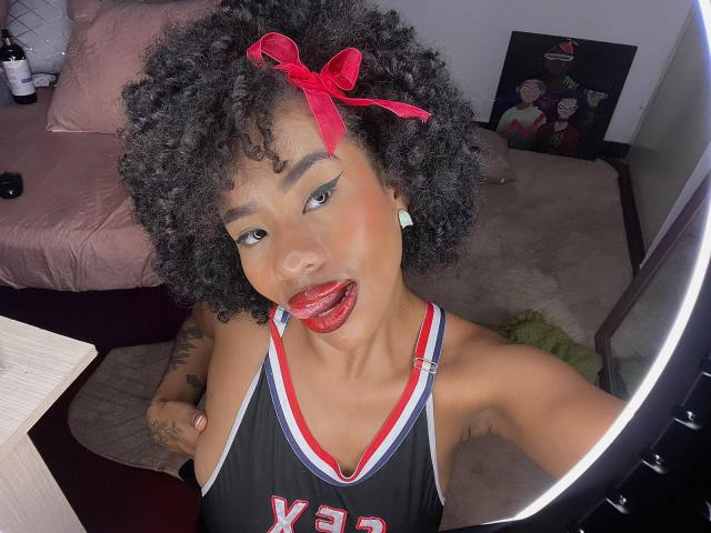 Karla, con i capelli neri ricci, un fiocco rosso, rossetto rosso, la lingua fuori, indossa una canotta da basket nera con la scritta "Varsity", riflessa in uno specchio, con un tatuaggio sul braccio destro..