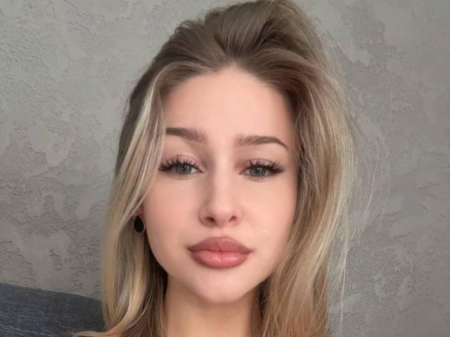 MilagrosEdmisten has blonde hair, green eyes, and full pink lips with black ear plugs. Her long eyelashes and slightly parted lips are visible. She's looking straight at the camera.