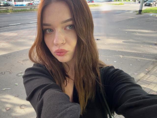 KiaraTease with puffy pink lips, green eyes, and brown hair in a black top, taking a selfie with a serious expression, showing her small tits and slim face.