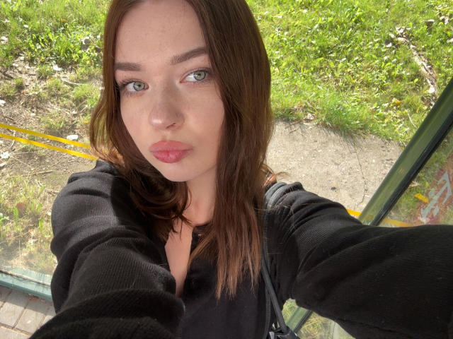 KiaraTease a les yeux verts, les lèvres roses et les cheveux bruns. Elle porte un pull noir et prend un selfie en extérieur, avec un fond jaune et vert. Ses seins ne sont pas visibles..