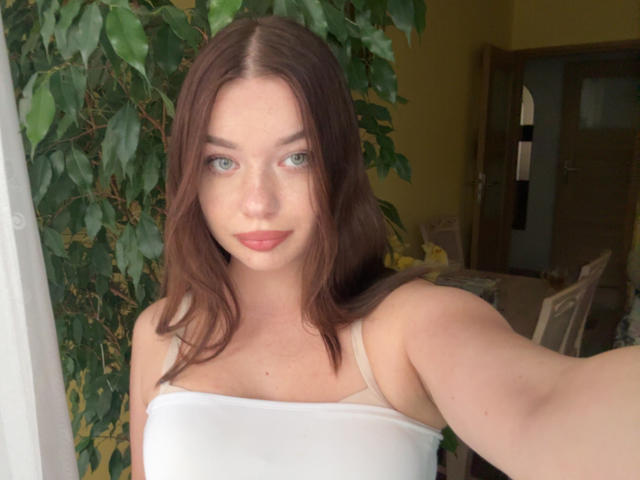 Kiara, avec ses cheveux bruns moyens, ses yeux bleus et sa peau pâle, porte un haut blanc sans bretelles, qui révèle ses petites seins et les bretelles légèrement visibles de son soutien-gorge bronzé..