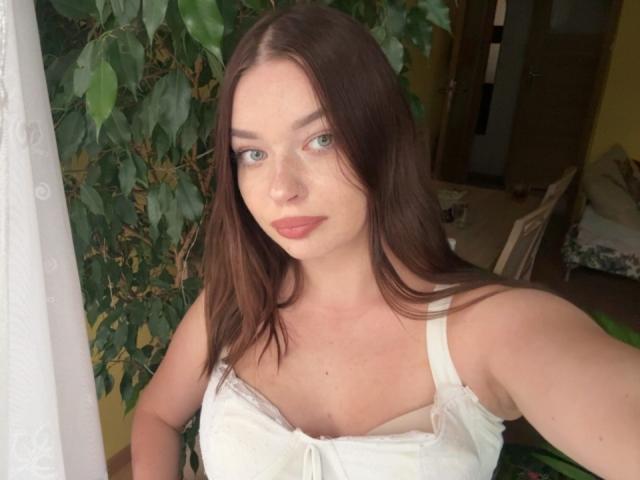 KiaraTease a la peau pâle, les yeux verts et de longs cheveux bruns. Elle porte une robe blanche qui laisse entrevoir ses petites seins, et se tient près d'une plante verte, en regardant l'appareil photo..
