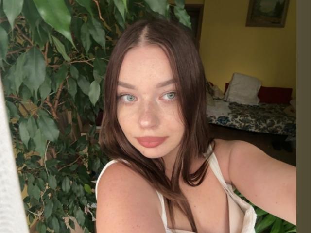 KiaraTease a les yeux verts clairs, les cheveux bruns et du rouge à lèvres. Elle porte un haut blanc qui dévoile ses seins, avec une plante à feuilles sur le côté gauche..