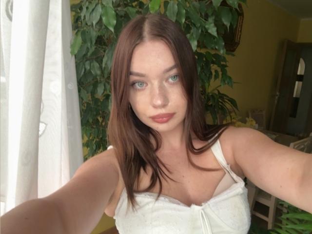 KiaraTease a la peau claire, les yeux verts et les cheveux bruns. Elle porte un haut en dentelle blanche qui révèle ses petites seins. Elle prend un selfie avec un regard calme et légèrement séduisant..