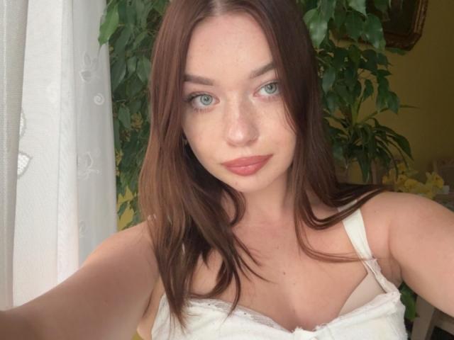 KiaraTease a les cheveux bruns, les yeux bleus, et porte un soutien-gorge en dentelle blanche qui révèle ses seins, avec un sourire discret et du rouge à lèvres rose. Sa peau est claire et lisse..