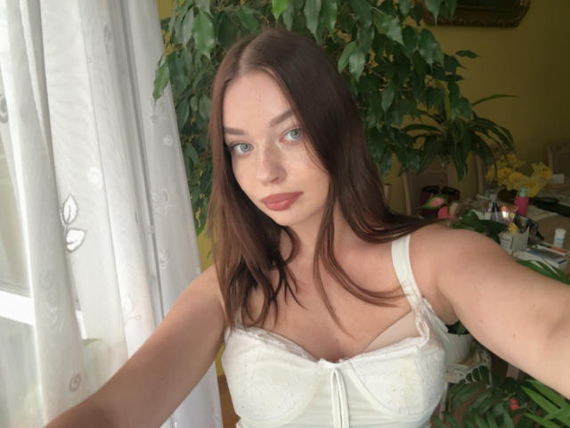Kiara, avec ses cheveux bruns moyens, ses yeux verts clairs et son rouge à lèvres rouge, porte un soutien-gorge en dentelle blanche et prend un selfie, montrant ses seins, dans une pièce lumineuse avec une plante..