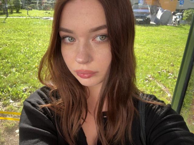 KiaraTease a les yeux vert clair, les cheveux bruns ondulés et les lèvres roses, elle porte un haut noir, avec une expression sérieuse, et se tient à l'extérieur, au soleil..