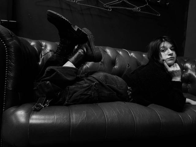 AlicaMihal lies on a tufted leather couch, legs up, wearing boots and jeans, black sweater, hand on cheek, black-and-white photo, sexy, relaxed, boots pointing upward.