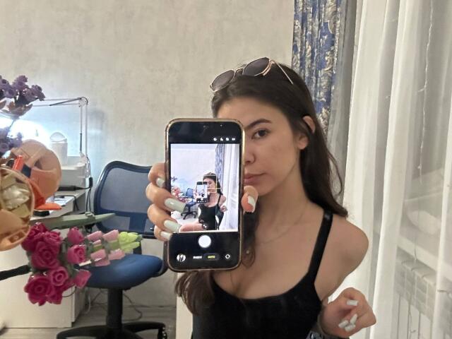 AnneKofron with long brown hair, wearing a black tank top, holding a phone in front of her face, showing her reflection with her small tits visible.