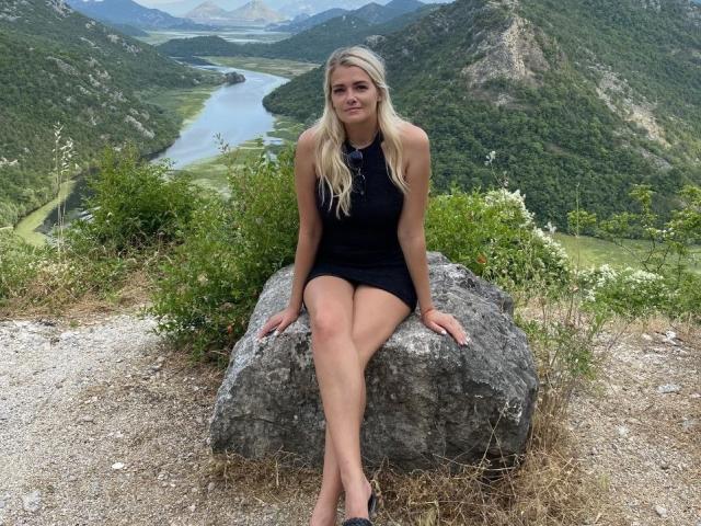 GladisBelle with long blonde hair, wearing a black sleeveless dress, sitting on a rock, showing her ass, and legs crossed, in a mountainous, green valley.