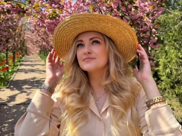 GladisBelle with blonde wavy hair, wearing a tan hat, beige shirt, and gold bracelet, smiling up, hands touching hat, blue eyes, and a cross necklace.