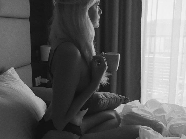 Black-and-white photo of GladisBelle with long blonde hair, holding a mug, sitting on a bed, knees up, arms around a pillow, wearing a tank top, looking out a window.