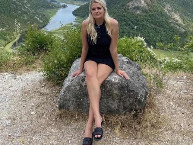 GladisBelle, blonde, black sleeveless dress, legs crossed, black peep-toe heels, sitting on rock, green mountains, river in background, grassy and rocky terrain, subtle smile, long hair.