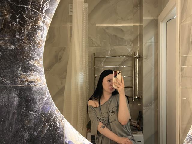 EvaTeasy with black hair, wearing a gray, off-shoulder top, taking a mirror selfie in a marble bathroom. She has a tattoo on her left forearm.