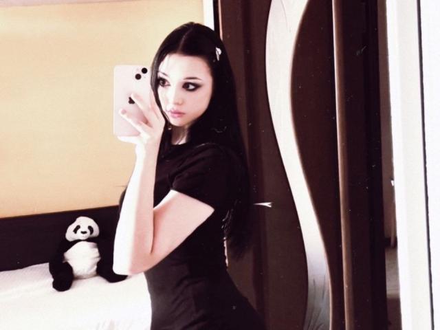 MaganSplonskowski with long black hair, wearing a black shirt, holding a pink phone, taking a mirror selfie, standing by a bed with a stuffed panda.
