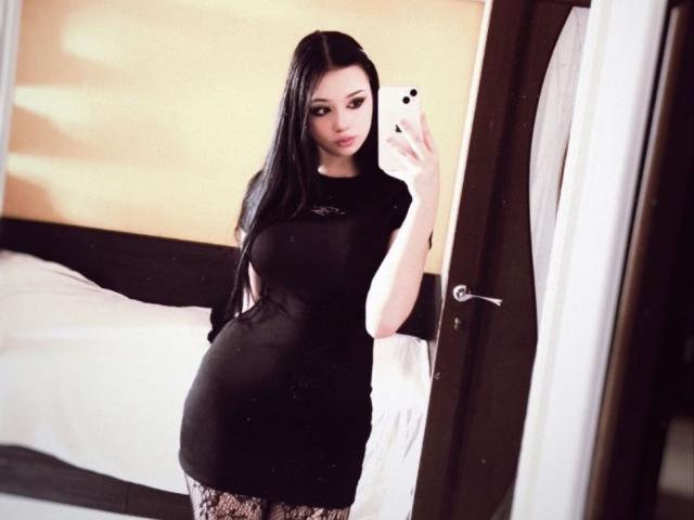 MaganSplonskowski with long black hair, wearing a tight black dress, taking a selfie in a mirror, showing a pregnant belly, and lace-patterned pants, holding a white phone.
