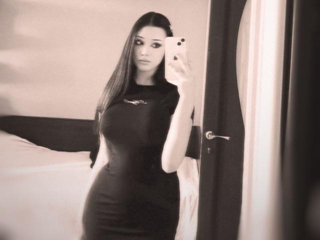 MaganSplonskowski with long dark hair, wearing a tight black dress, taking a selfie in a mirror, showing her big tits and curvy ass, standing in a softly lit room.