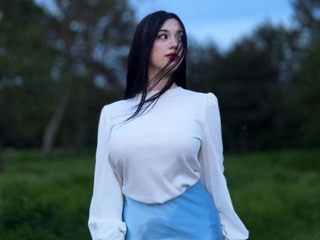 MaganSplonskowski with long black hair, white blouse, and blue skirt, standing outdoors, looking to the side, showing her tits and ass slightly visible under her blouse.
