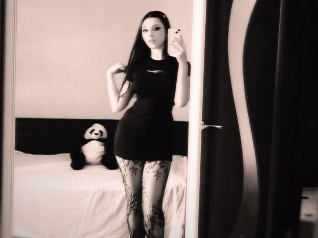 MaganSplonskowski with long black hair, wearing a tight black dress, lace stockings, and holding a phone, standing in front of a mirror, with a panda stuffed toy behind her.
