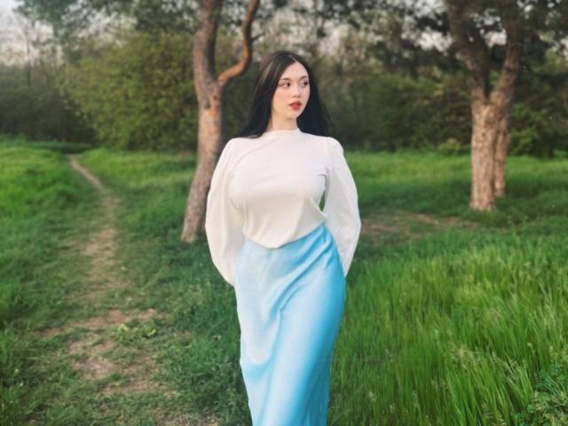 MaganSplonskowski with long black hair, white long-sleeve top, and blue high-waisted skirt, standing in grassy area, hands in pockets, trees in background, looking slightly to the side.